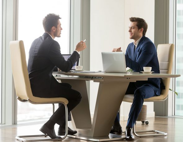 Two handsome businessmen sitting in comfortable chairs at desk with laptops in meeting room. CEO making important negotiation about companies partnership or corporate merger. Financiers planning deal